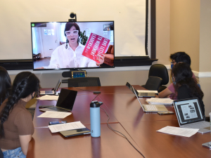 Class room with students around a large table interacting with a virtual guest speaker on a screen