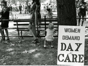 B & W image of two women and a baby in a city park with others in the background with a sign "Women Demand Day Care"