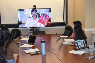 Class room with students around a large table interacting with a virtual guest speaker on a screen