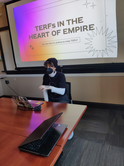 Emma sits at a desk giving a presentation. The screen behind her reads "TERFs in the Heart of Empire"p